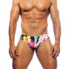 Men's swimwear, slip style, worn by a man, front view, tropical pattern with green vegetation base and various types of pink and yellow flowers.